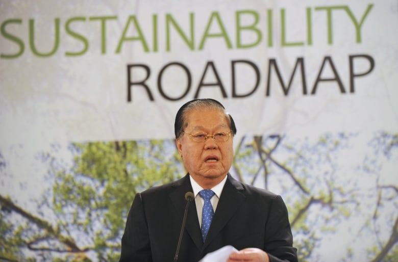 A man wearing a dark suit, white shirt, blue tie and glasses speaks into a small microphone. Behind him is a large poster with a photo of a tree and the words 'Sustainability Roadmap.'