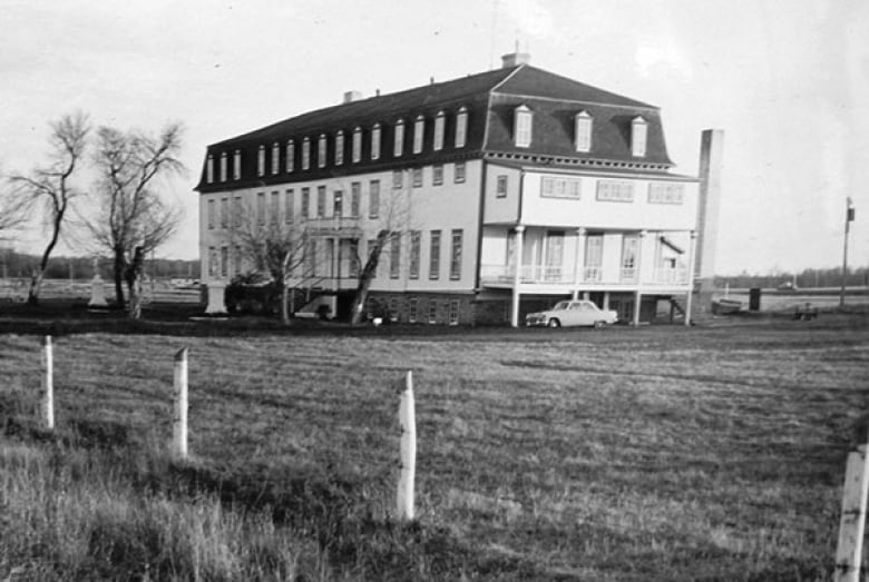 Older black-and-white photo of a building in a field with a few trees and a car out front.