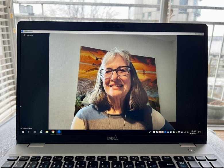 A middle-aged white woman sitting in front of a painting is seen on a laptop screen during a Zoom call. 