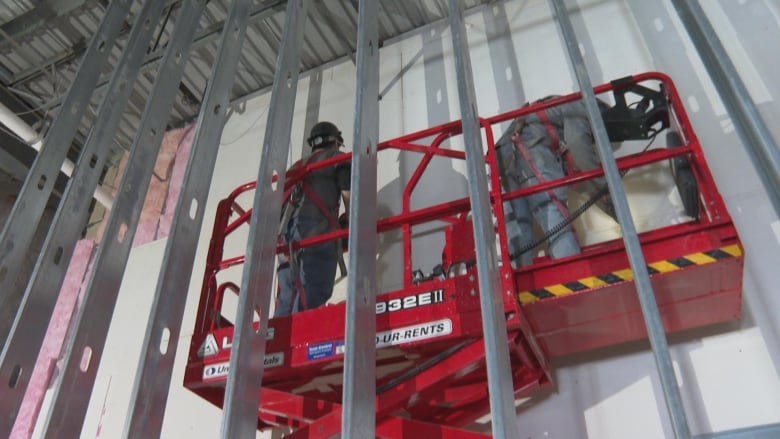 Two construction workers stand on a red scissor lift installing wall boards in a building under construction.