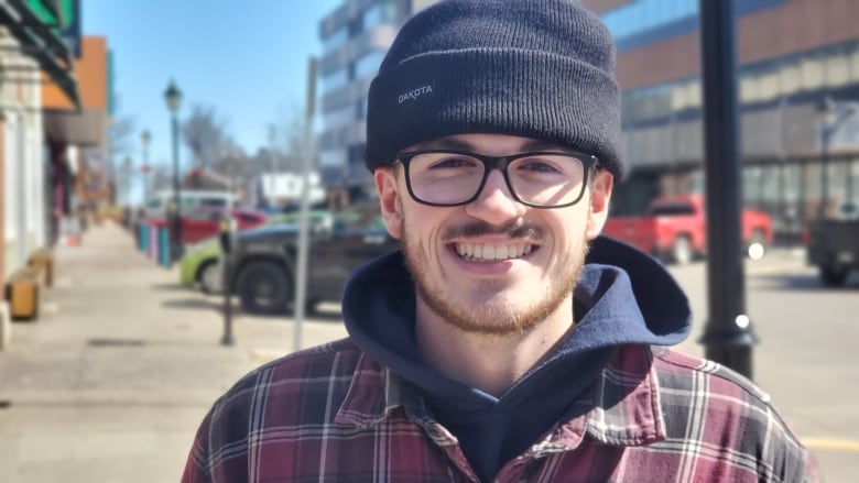 A man wearing black-rimmed glasses and a dark blue toque smiles at the camera while standing on a sidewalk downtown. 