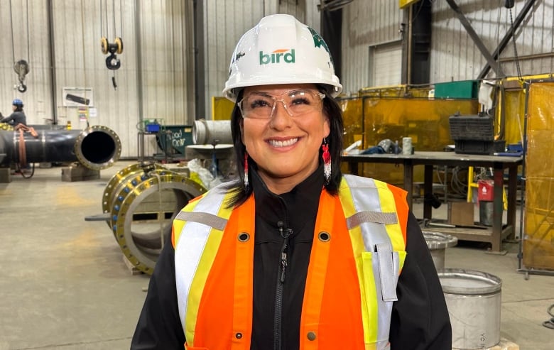 A smiling woman in a white hardhat bearing the Bird Construction logo, safety goggles and a orange and yellow safety vest stands in an industrial fabrication shop. 