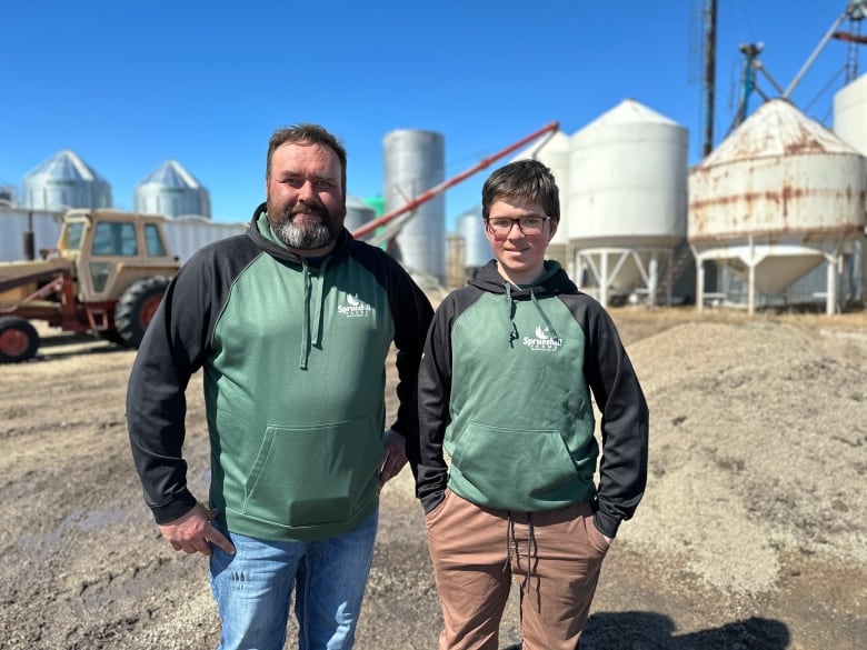 A man in his 40s stands next to a teenager look at the camera on a sunny day. They wear matching hoodies with the family farm logo on the chest. 