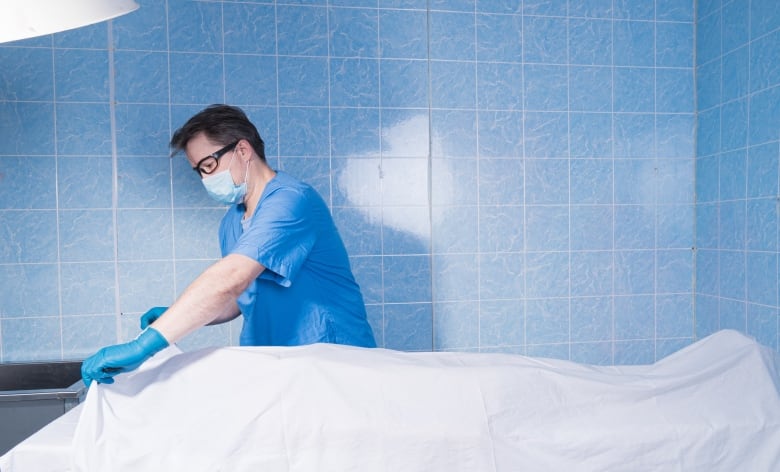 A white man wearing dark-framed glasses or goggles, a white surgical mask, blue short-sleeved scrubs and blue plastic glasses lifts a white sheet from a body lying on a gurney.