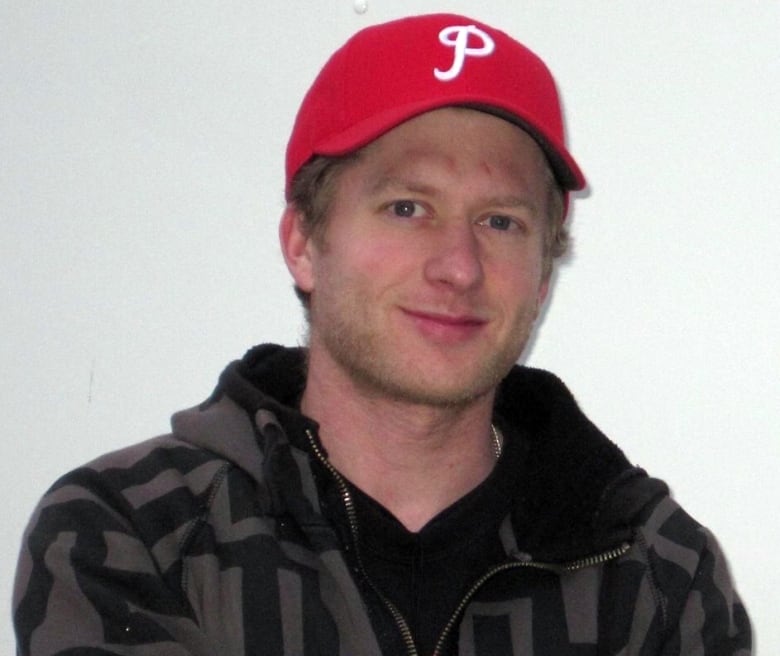 A young white man with a red baseball cap and grey and black patterned hoodie is giving a slight smile in front of a white background.