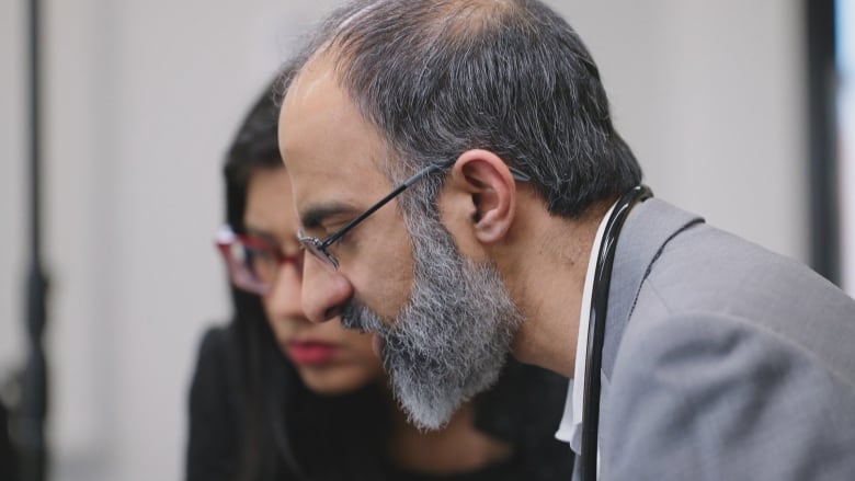 The profile of a man with a grey beard, thinning grey hair and glasses is shown in centre frame. He appears to be looking down at something to the left of frame. A woman with dark hair, red glasses and red lipsticks appears out of focus just to the left of him, looking in the same direction.