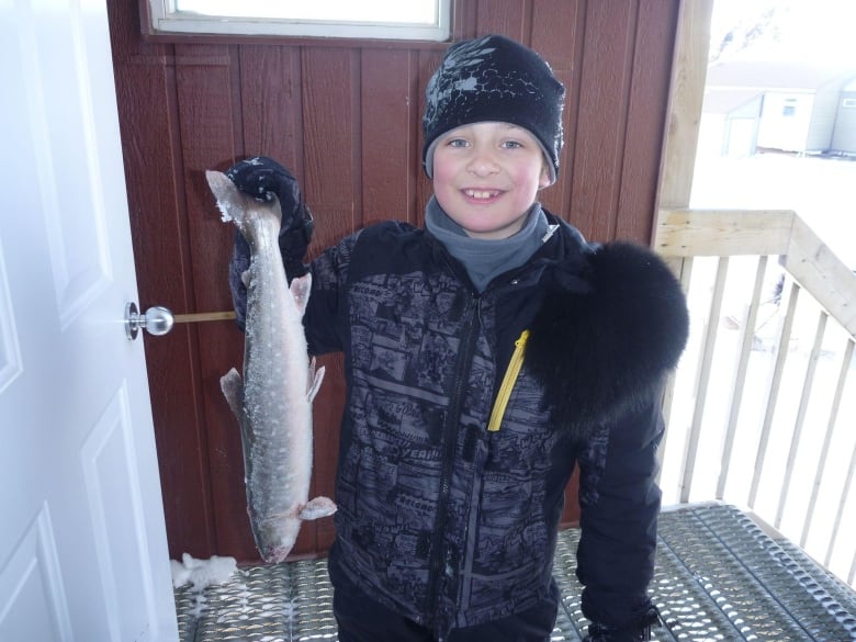 A kid stands outside in winter outdoor clothes holding a fish 