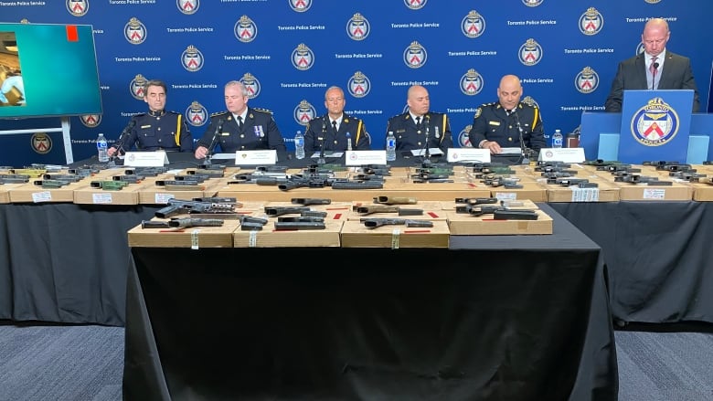 People sitting behind a table of guns. 