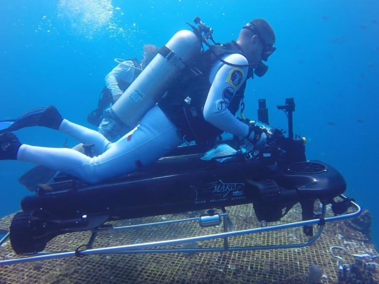 Canadian astronaut Jeremy Hansen is in a grey diving suit in the ocean sitting atop a small black vehicle that propels him through the water.