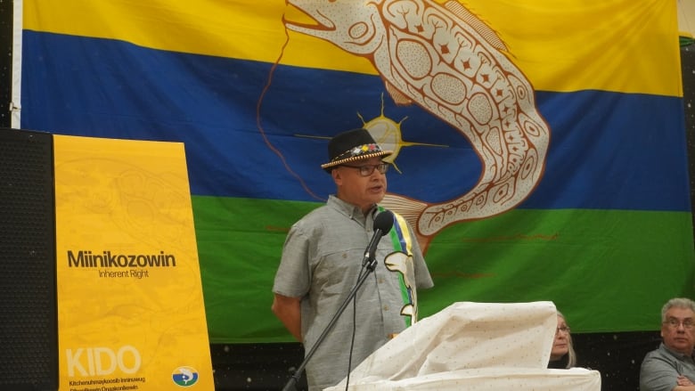 A man stands at a podium with a First Nation flag behind him.