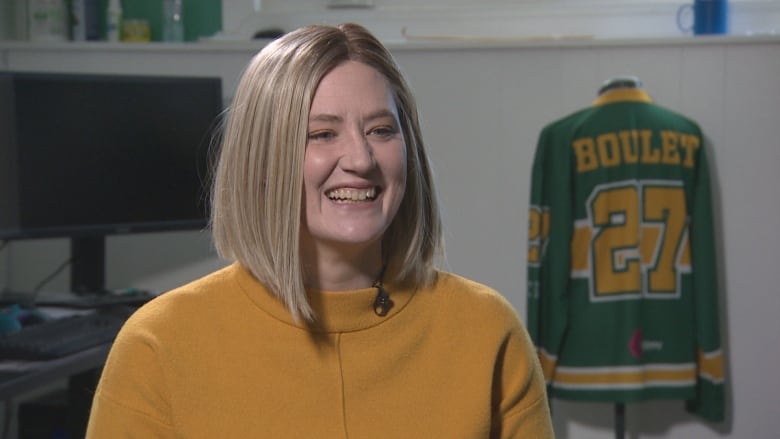 Brandy Hehn in a yellow shirt smiles while sitting in front of a Green Shirt Day jersey.