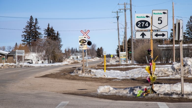 Road signs and a small memorial of flowers and hockey sticks are pictured along a highway lined with snow. 