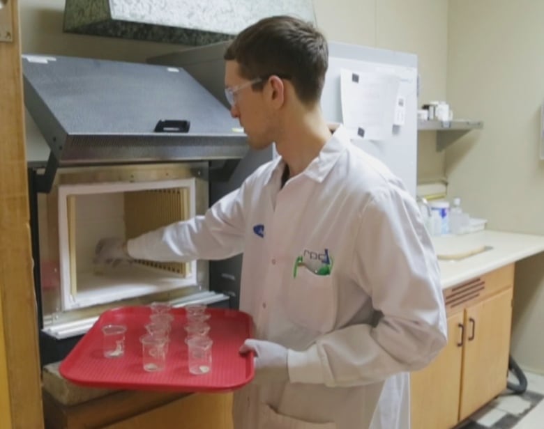 A man in a lab coat holding a red tray of small beakers and placing one of them inside an open metal box