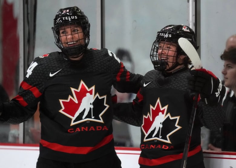 Two female hockey player smile while standing side by side with their arms around each other's back.