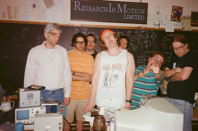 Seven men pose in a grainy photo. Behind them is a blackboard with mathematical formulas on it. Above is a sign that says "Research in Motion."