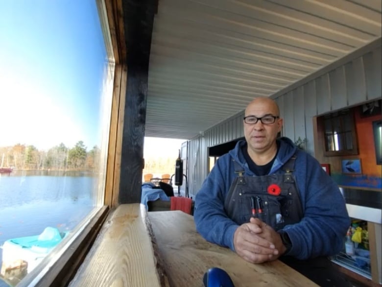 A photo of Joe Nimens sitting at a table inside his converted shipping container floating home, beside a window overlooking a lake. 