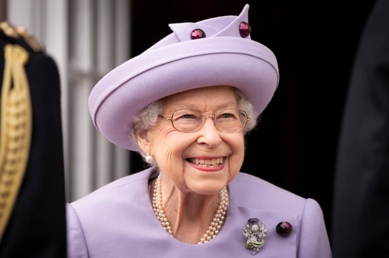 Grey haired woman with glasses, wearing lavender coat and hat, smiling