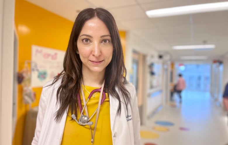 A doctor with long brown hair, wearing a white lab coat and a yellow shirt, with a stethoscope around her neck, stands in a hospital hallway.
