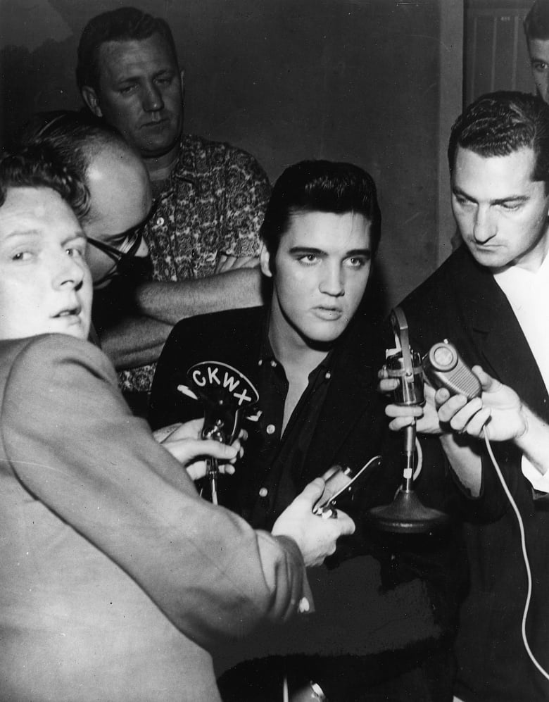 A picture of Elvis Presley being interviewed by journalists with Red Robinson in the foreground.
