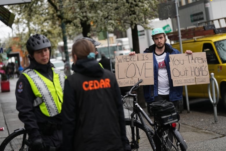 A police officer talks to a person wearing a jacket marked 'CEDAR', while a person in the background holds up two signs reading 'Where is Love' and 'You Threw out HOMES'.