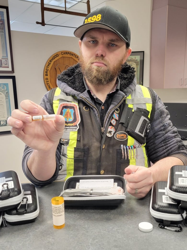 A man in a black cap and yellow high visibility vest sits behind a counter with naloxone kits on it and shows the contents of one of the kits.