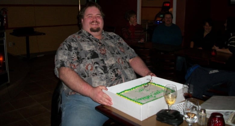 Obese man sitting at a table with a birthday cake in front of him. 