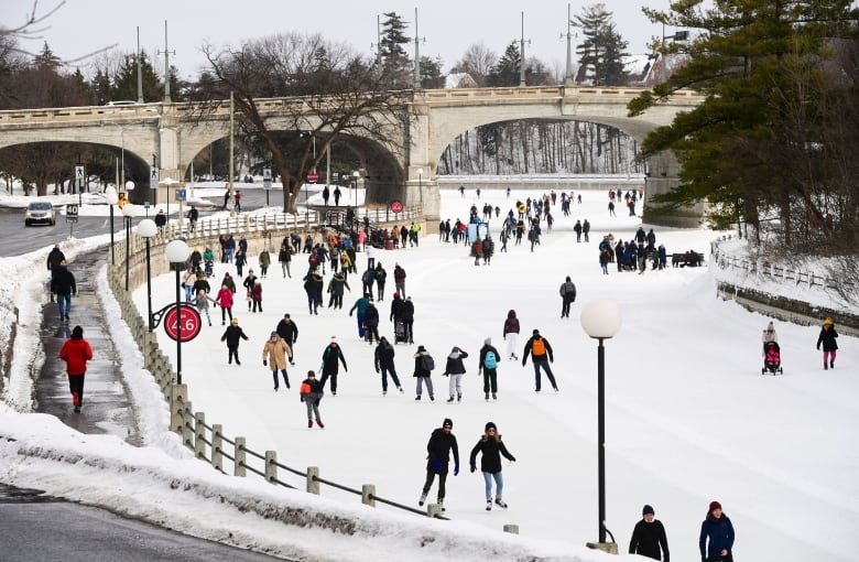Many people skate on a frozen canal near a road and bridge.