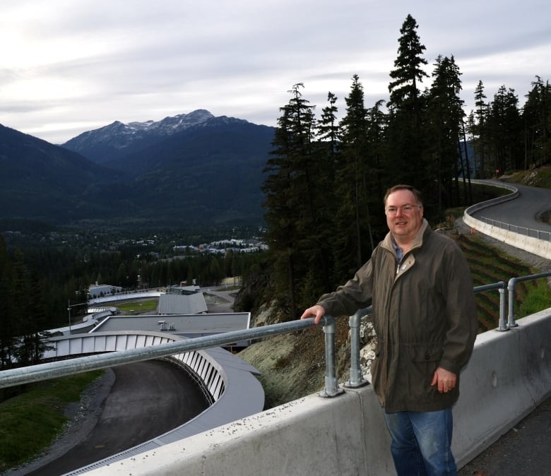A man in a green coat stands near a luge run in the mountains.