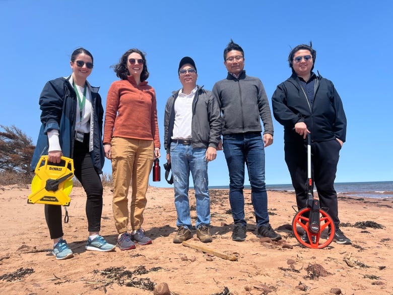 Five people stand on a beach, three of them carrying tape measures and other measuring tools. 