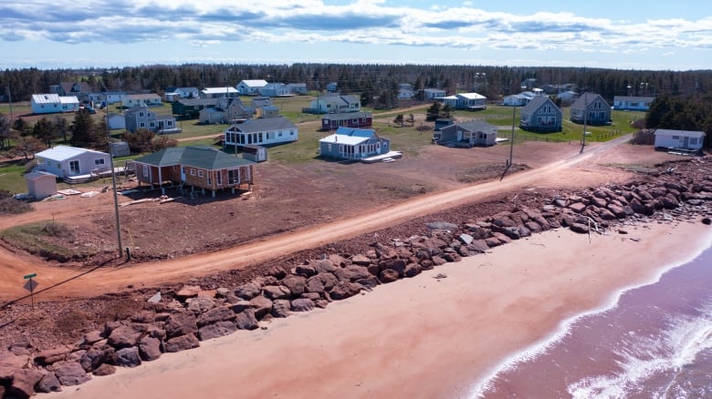 Drone shot shows a cottage subdivision near a beach that has large sandstone blocks piled against the bank to keep it from eroding.