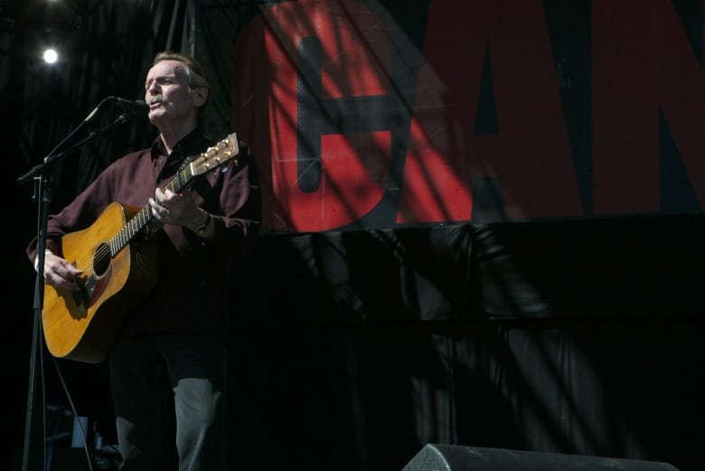 Holding a guitar, a man sings in front of a microphone on stage during a benefit concert.