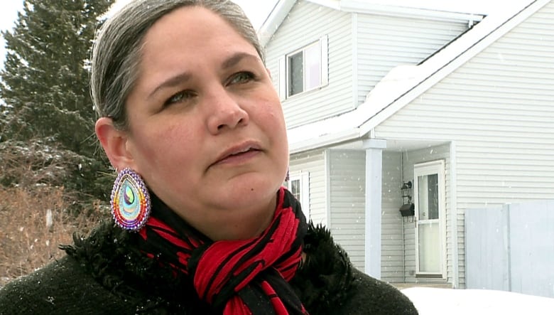 A woman stands in front of her home on a snowy day. She is wearing a red scarf and colourful earrings. 