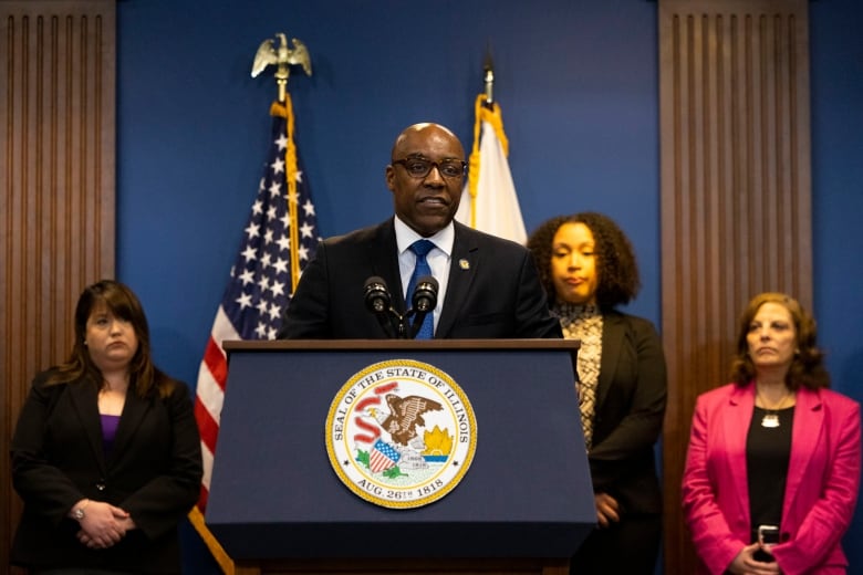A Black man standing at a podium with the seal of the State of Illinois on it. He's wearing a black suit with a blue tie. There are three people standing behind him.