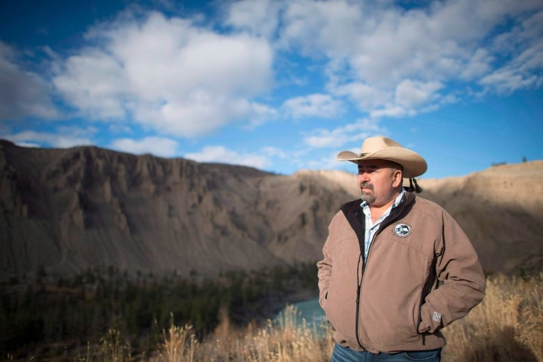 An Indigenous man wearing a cowboy hat poses in front of a hill.