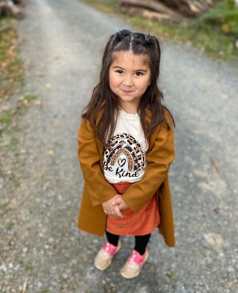 A young girl with brown hair and brown eyes smiles for the camera. Her hair is in pigtails and she wears a burnt orange cardigan. 