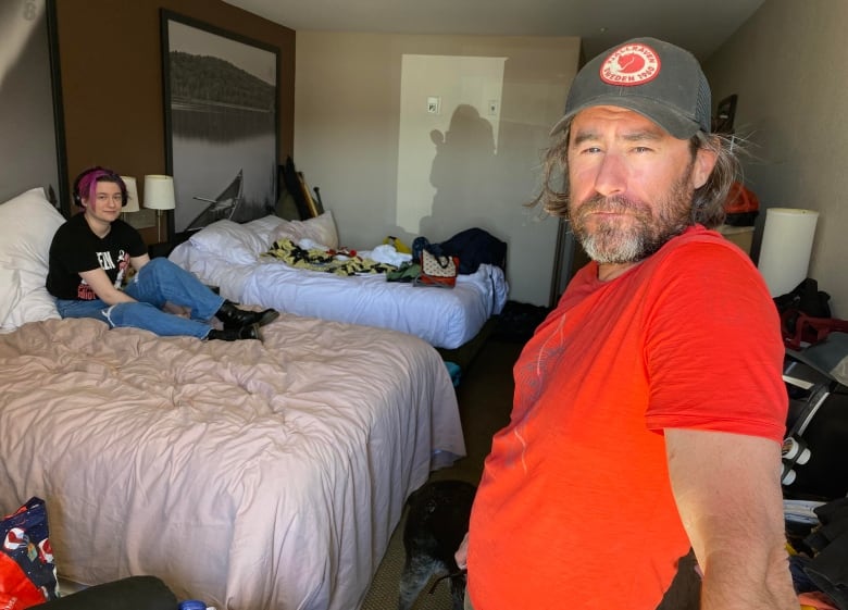 A man wearing a t-shirt and a baseball hat stands in the doorway of a hotel room. Behind him, his teenage daughter sits on a bed with various belongings on the other bed and on the floor.