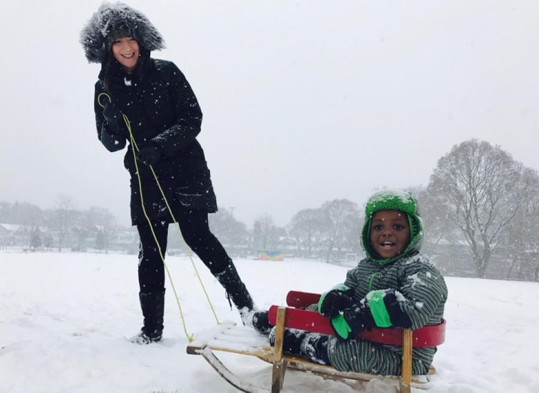 A smiling little boy sits in a snow sled while his mother pulls it. It is snowy. 