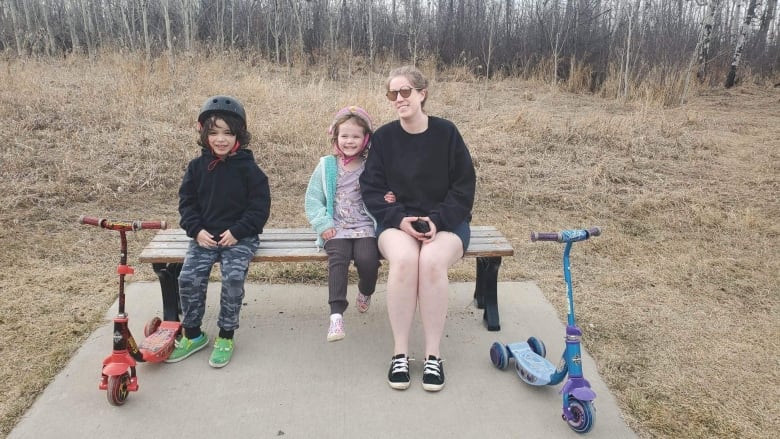 A woman wearing a black sweatshirt and shorts sits on a park bench with two children sitting beside scootters and wearing brightly collared bike helmets. 