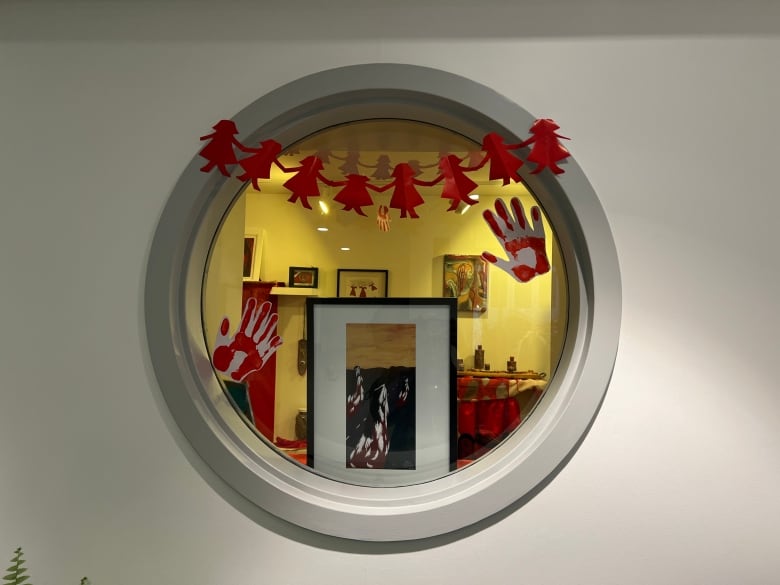 Photo of circular window with paper dolls, paper red hands and paintings in the background.