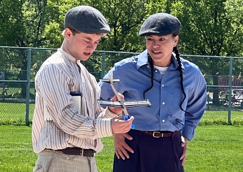 Two actors wearing newsboy caps, button-down shirts and belted baggy dress pants talk while performing outdoors in a city park.