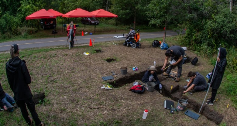 A wide shot of several people at an archaeological dig site. The site is surrounded by trees and is next to a bike path. 