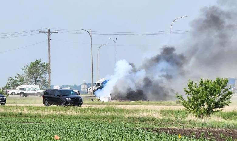 A large plume of smoke can be seen beside a highway, with a semi truck and emergency response vehicles in the background.