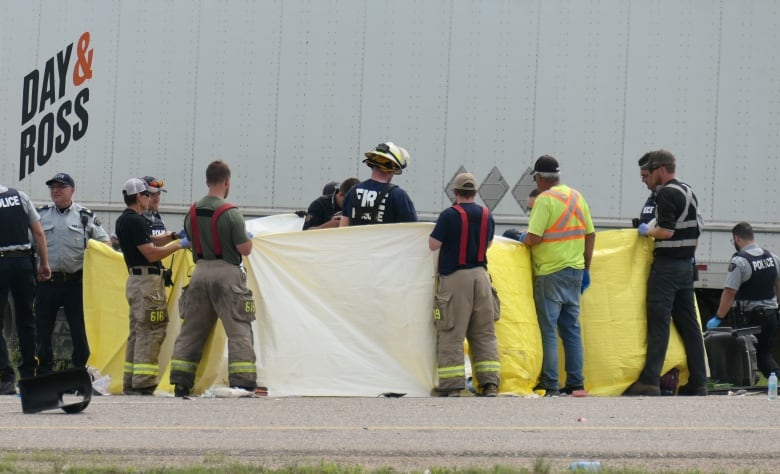 A group of police and firefighters at the scene of a highway crash hold up yellow tarps near a semi-trailer truck.
