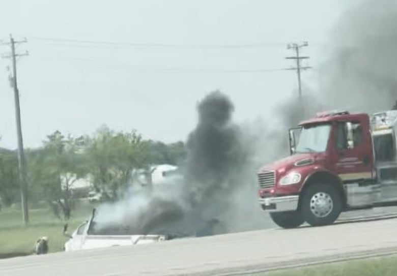 Smoke billows from a vehicle in a ditch. A semi-trailer truck sits nearby.
