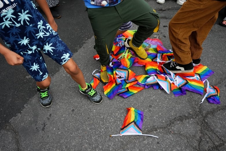Three kids stomp on a pile of small Pride flags on a road.