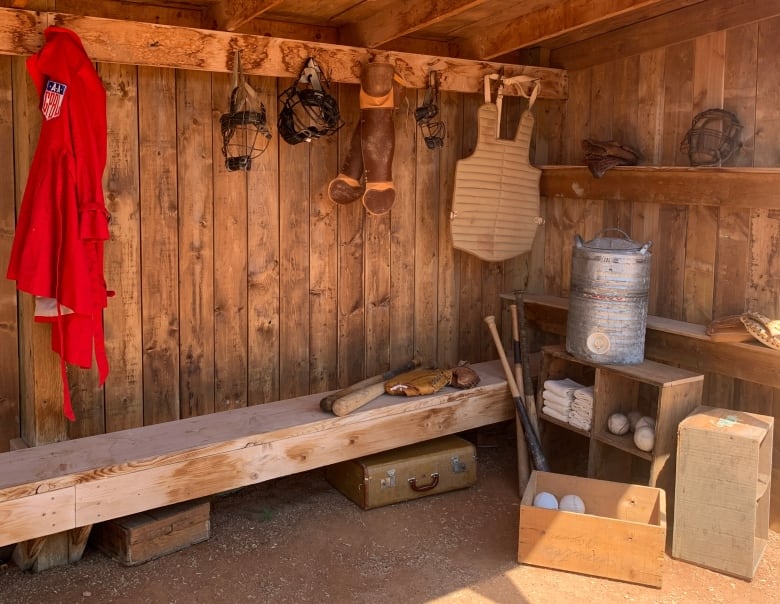  A baseball dugout with crates of balls, gear hanging from hooks and bats all decorating the inside. 