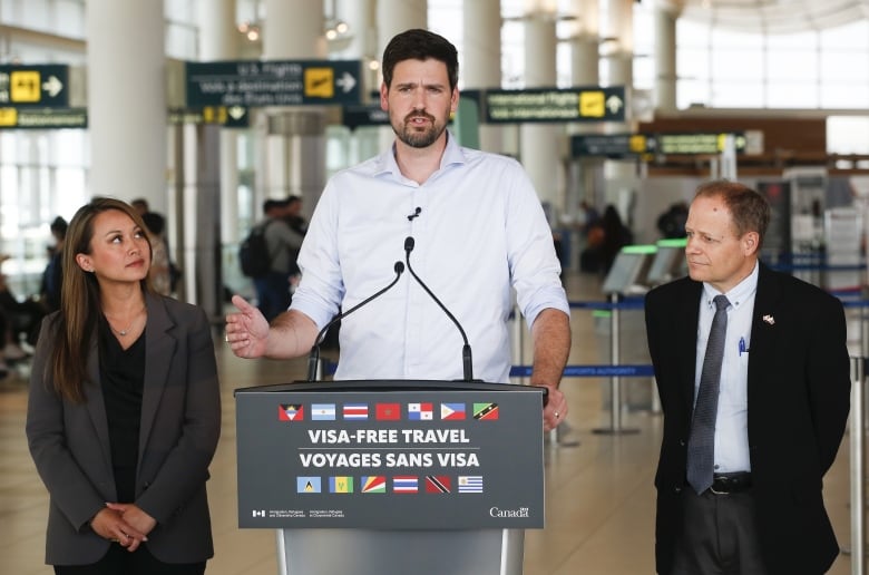 A man with dark hair and a beard wearing a white button-down shirt gestures while standing in front of a podium that reads 'visa-free travel' in English and French, as a man and woman wearing suits watch him speak. 