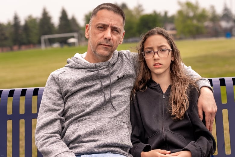A father sits on a park bench with his arm around his teen daughter's shoulder.