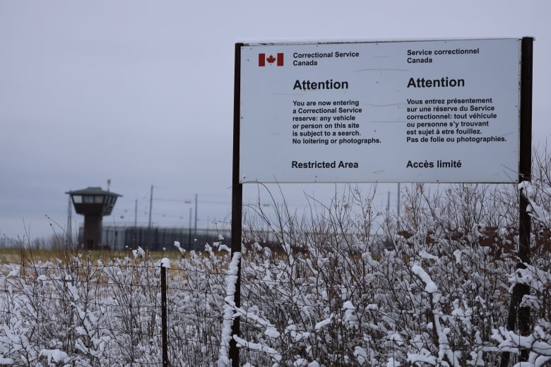 A sign is seen above a snowy fence outside a prison.