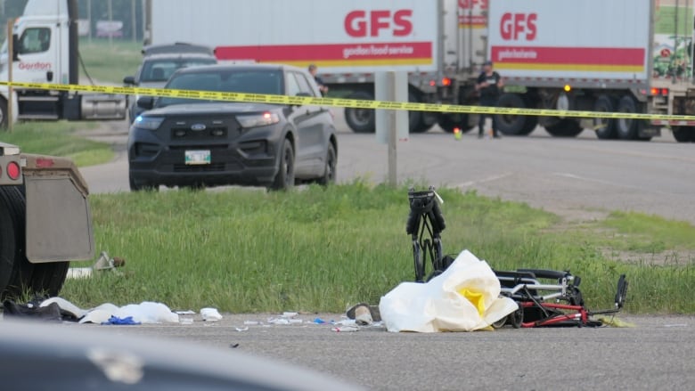 Two white and red semi-trailers are parked in the background while yellow police tape crosses the centre of the photo. A pair of folding walkers lie near the edge of a road. 